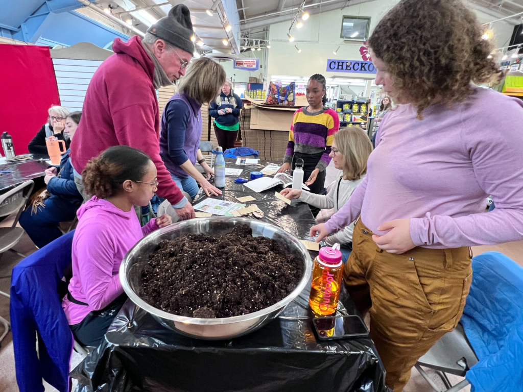 Big bowl of soil on a table in front of a gorup of people in various stages of picking out seeds they want to use