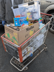 A shopping cart used by a food bank to transport food is overflowing with various food and hygiene items. A food bank worker stands behind the cart.